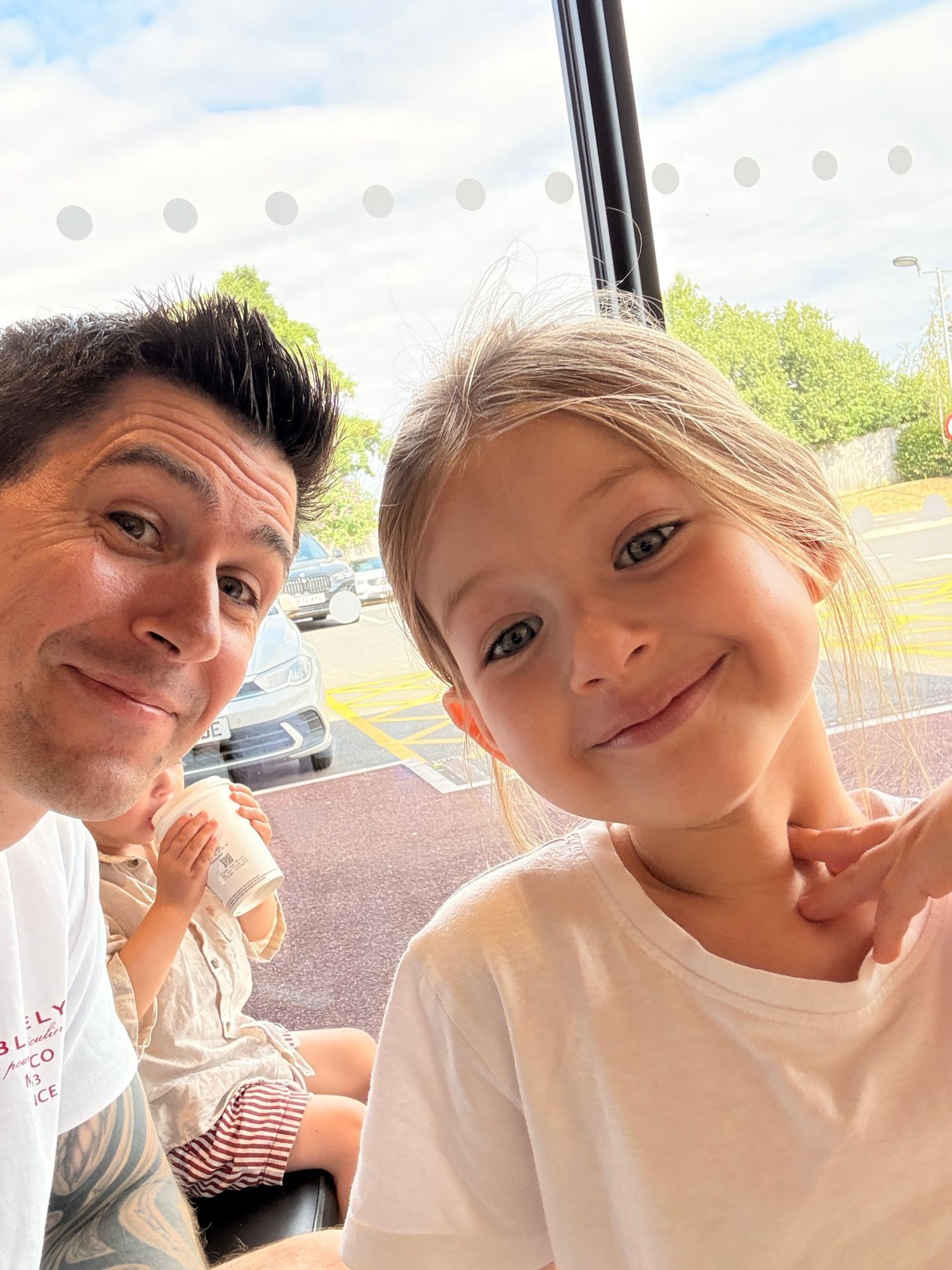 A selfie of me, Nate, with my daughter, both smiling in a café window, with my youngest drinking from a cup behind us.